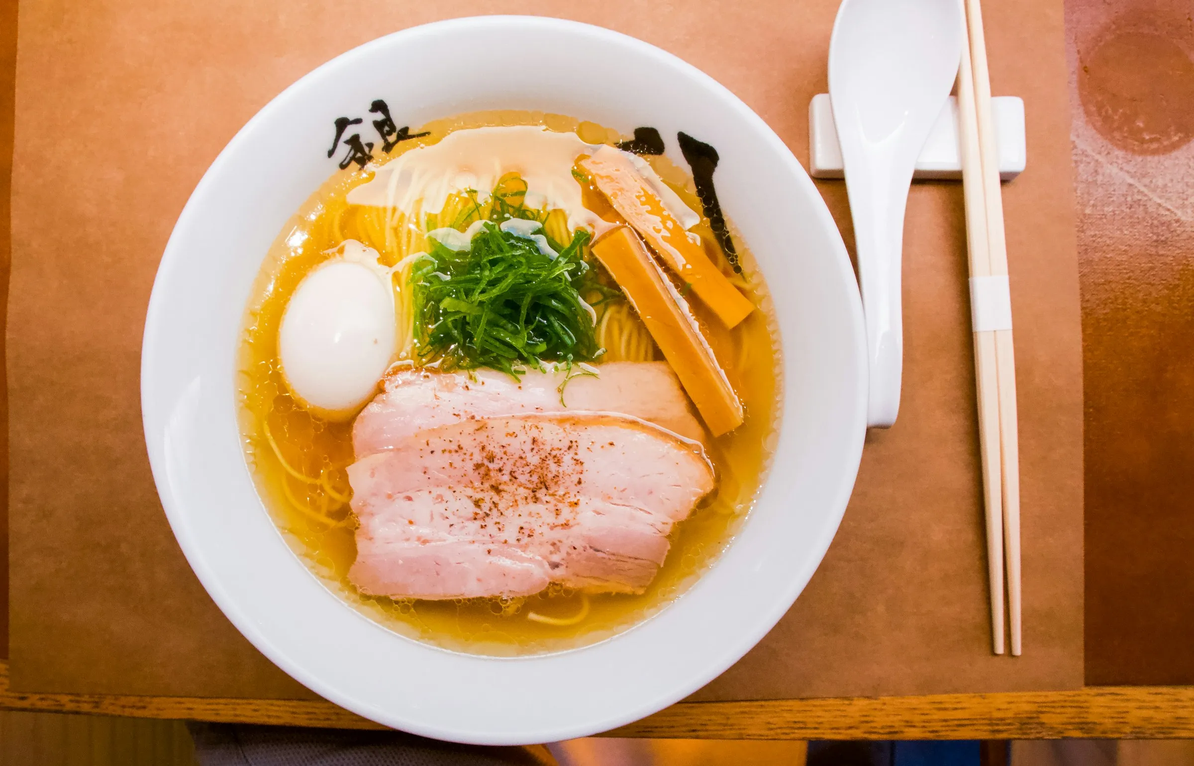 Ramen von oben mit Schweinebauch, Ei, Bambussprossen und Frühlingszwiebel, daneben Stäbchen und Löffel